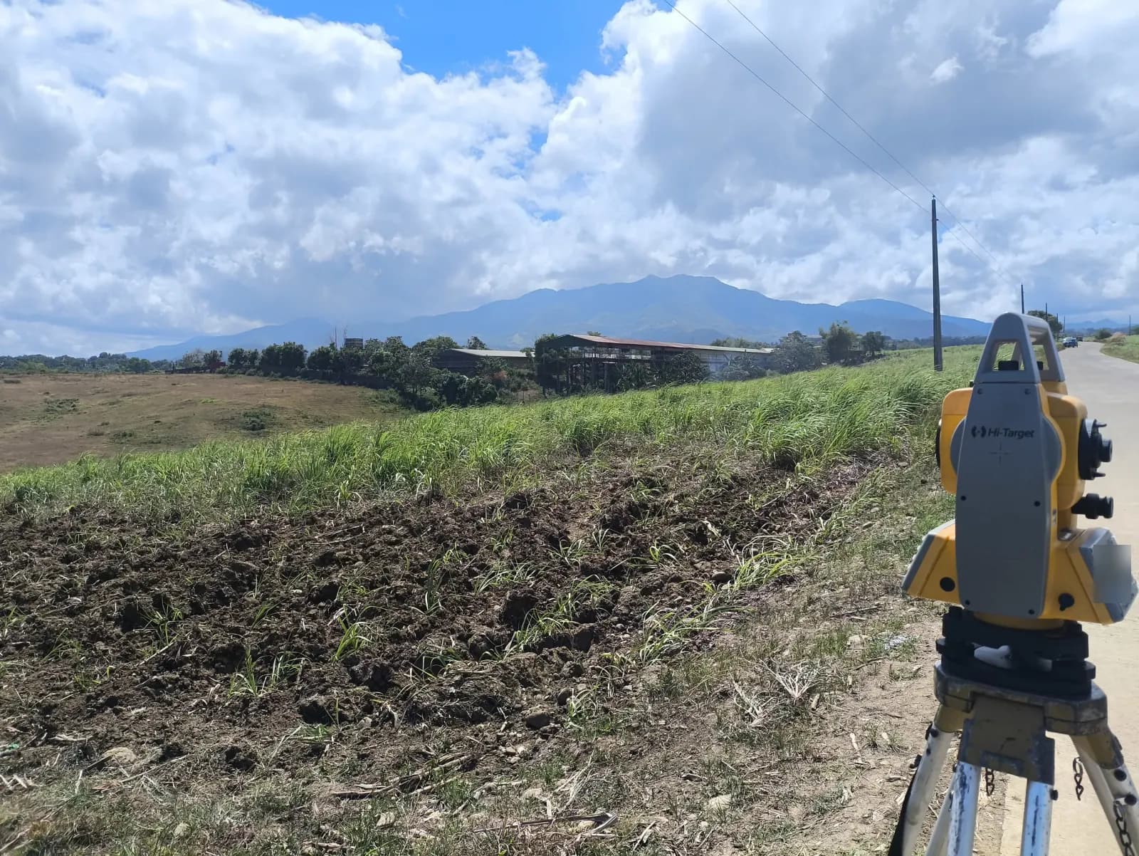 Total station beside a paved road and grassy verge, with tilled soil, utility poles, a long roofed structure, and a mountain ridge in the distance