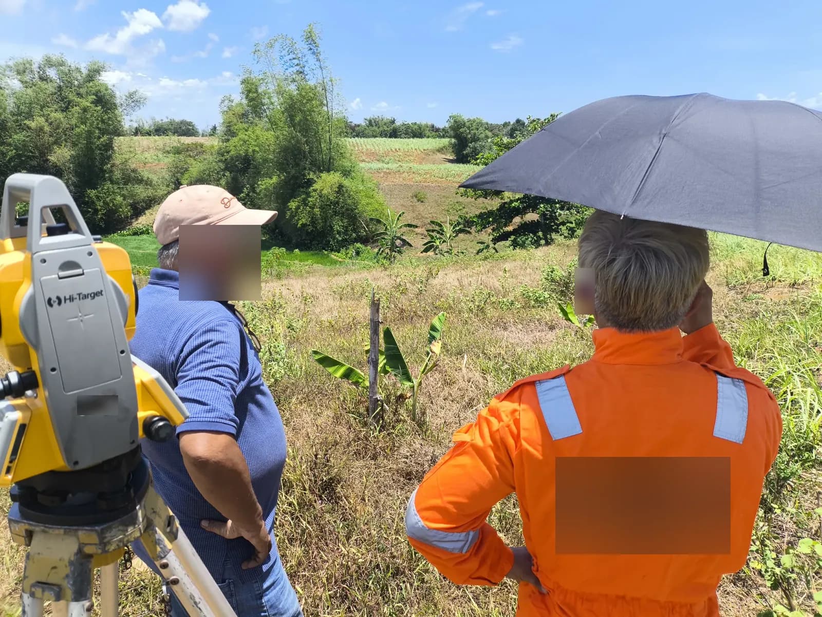 Two people beside a total station overlooking sloping farmland, one holding an umbrella, with banana plants, bamboo, and cultivated ground below