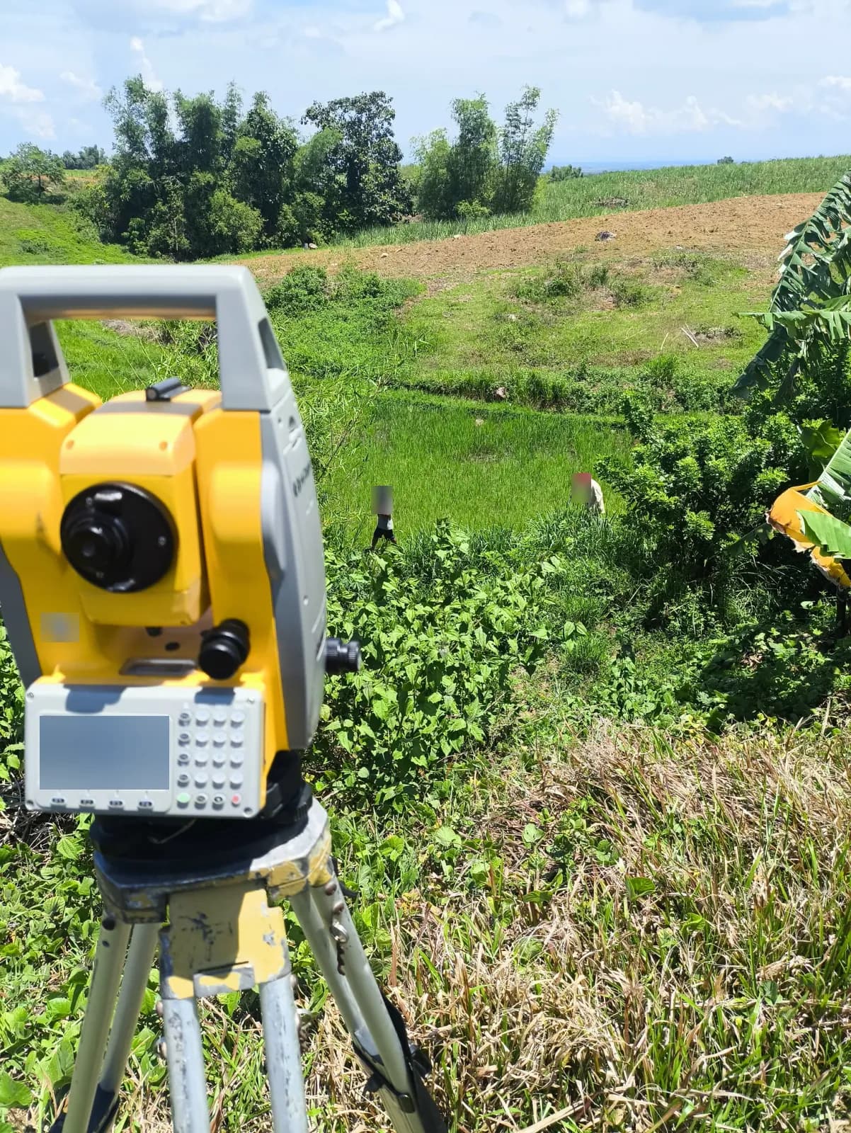 Total station in the foreground above a green field, with two people among the lower vegetation, banana leaves at the right, and cultivated slopes beyond