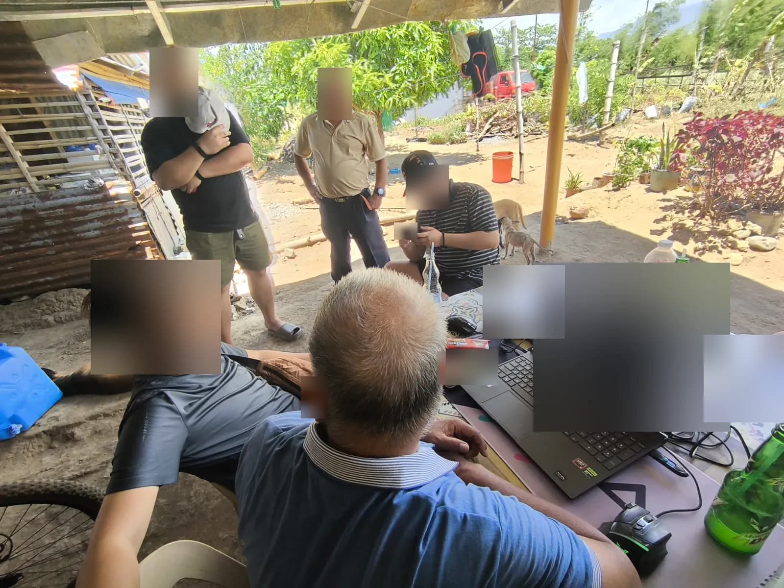 People gathered around a table under an outdoor shelter, with a laptop and paper records in front of them and a tree-lined yard beyond
