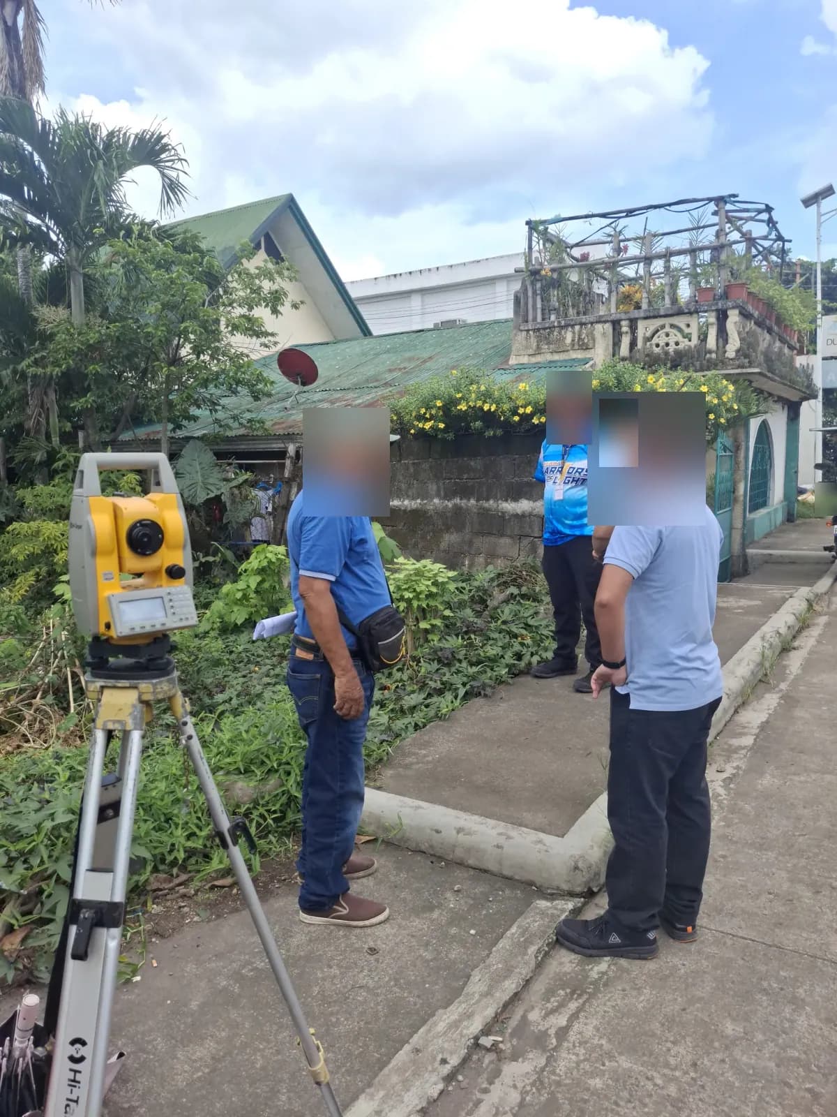 People gathered beside a total station on the sidewalk at the edge of the vegetated property
