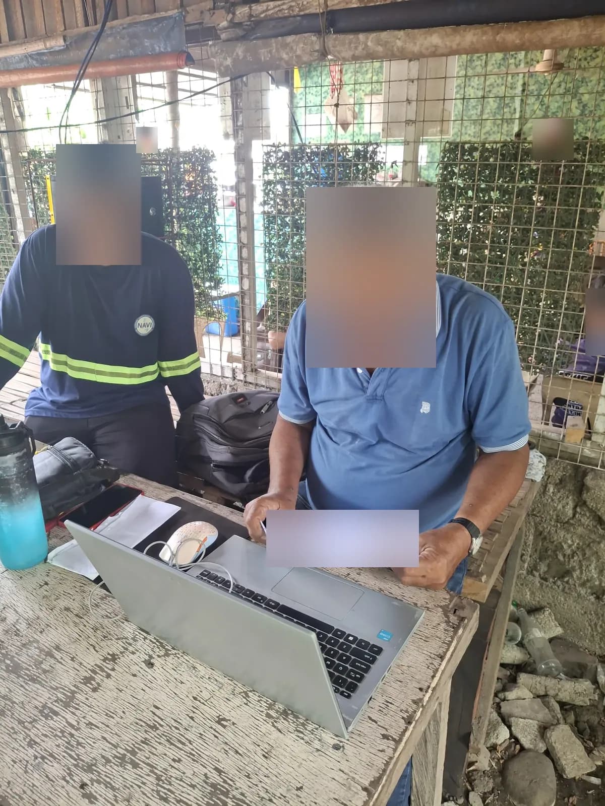 Two crew members reviewing printed documents with a laptop on a wooden table under a shelter