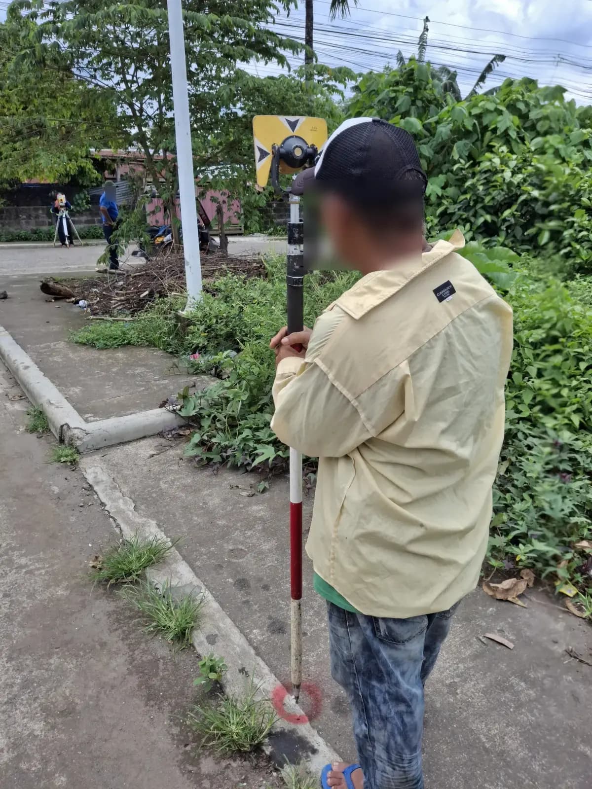 Rear view of a prism-pole holder at a red-painted curb mark with a total station across the street