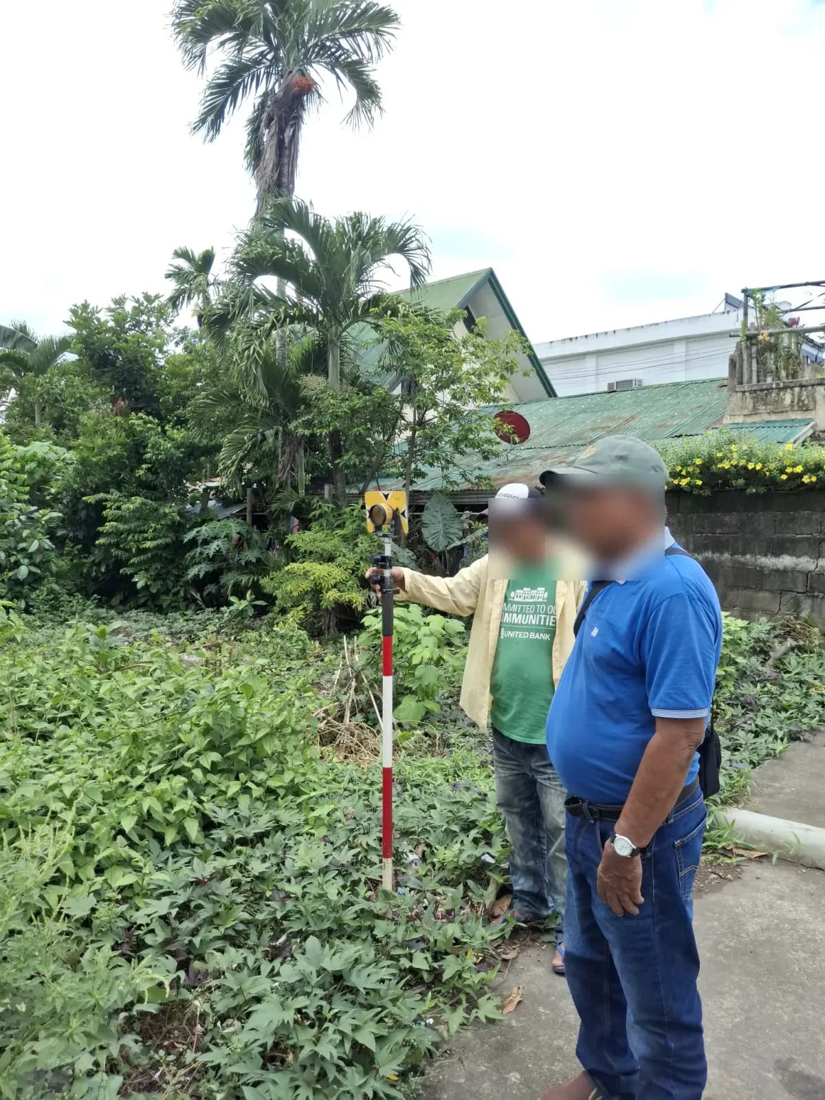 Two people beside a survey prism pole at the edge of a vegetated roadside lot