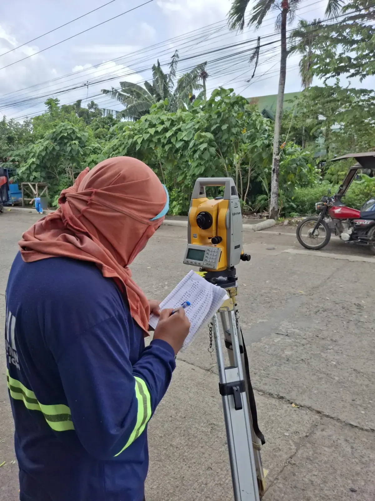 Survey operator recording field notes beside a tripod-mounted total station on a neighborhood street