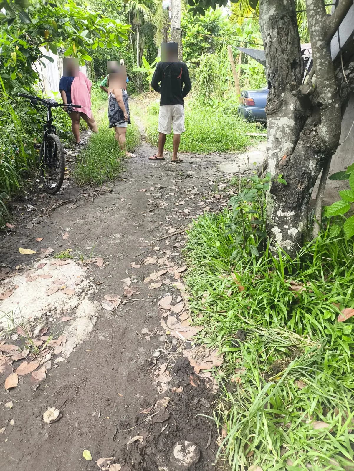 People gathered on a narrow dirt path between homes and vegetation, with a bicycle and parked car nearby