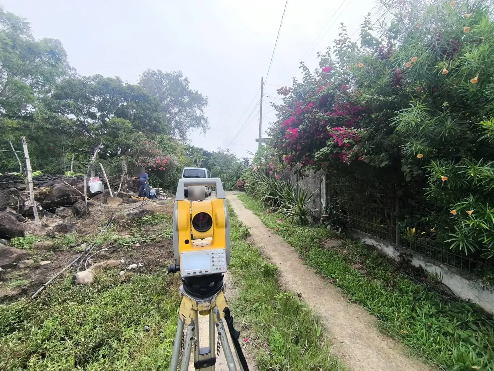 Front view of a total station beside a narrow concrete path, with a rocky fenced site on the left and a flowering wall on the right