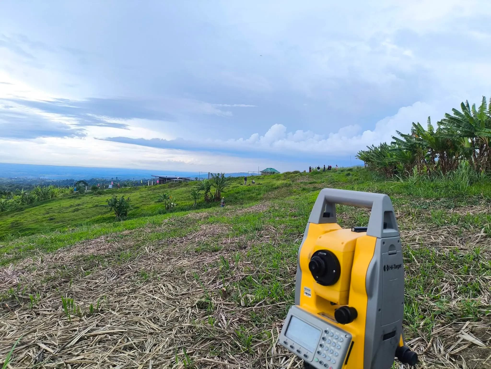 Leonor Surveying Services Hi-Target yellow total station on a tripod positioned on an elevated hilltop in Silay, Negros Occidental, with a wide panoramic view of the lowlands, clients gathering in the background, and dramatic cloudy skies during a parcelary survey