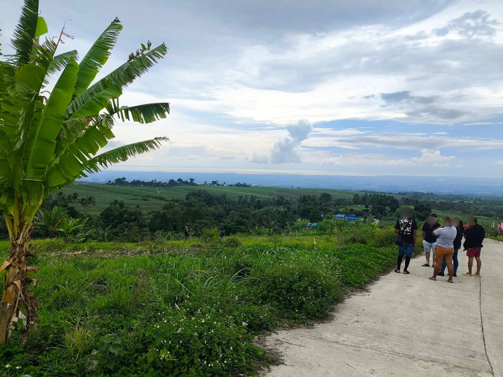 Panoramic hilltop view of the parcelary survey site in Silay, Negros Occidental, showing lush green agricultural valleys, sugarcane fields, a concrete farm road, a banana tree, and a group of blurred stakeholders reviewing the surveyed area with Leonor Surveying Services