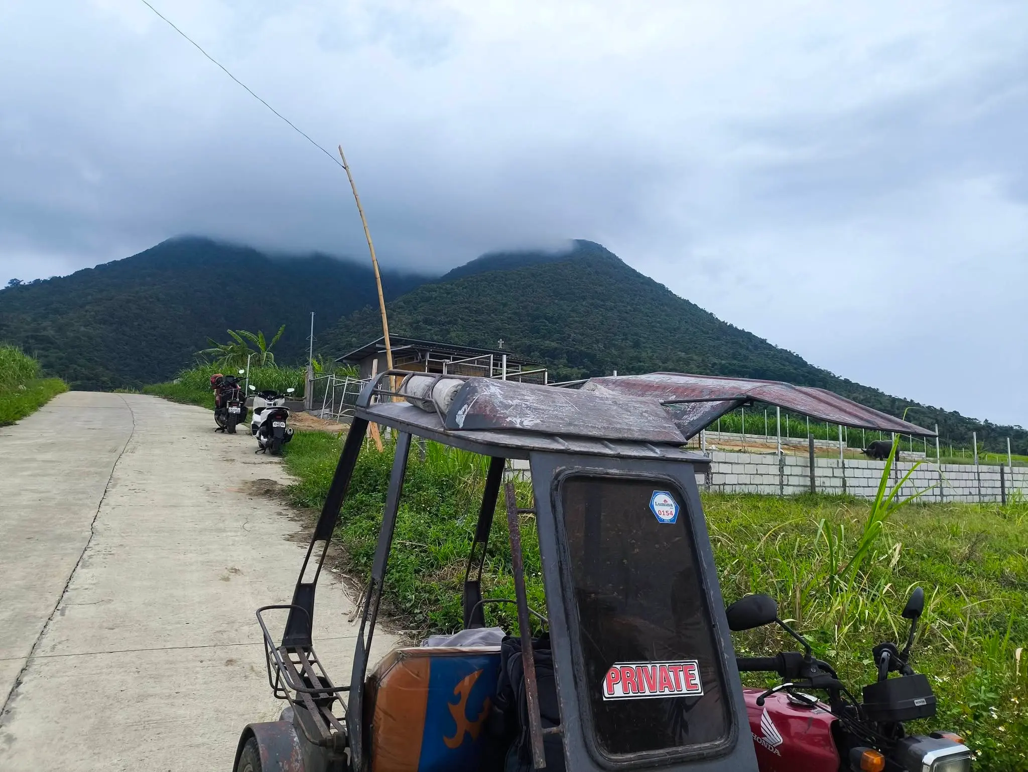 A private tricycle parked on a concrete road near the Leonor Surveying Services parcelary survey site in Silay, Negros Occidental, with dramatic cloud-covered twin mountain peaks rising in the background and a farm fence and small building visible along the road